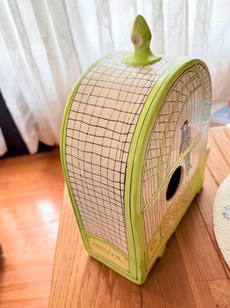 May include: A decorative birdhouse with a cage design. The birdhouse is painted in shades of green and cream, with a black grid pattern and a small bird illustration. The word "maison" is written on the front.