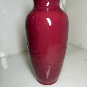 May include: A tall, glossy red vase with a slightly flared rim. The vase has a smooth surface with subtle embossed floral patterns. The vase is a deep, rich red color and is suitable for displaying flowers or as a decorative item.