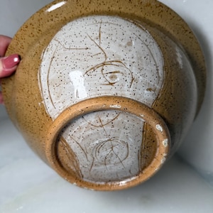 Peut inclure: Un saladier en céramique, vu de dessous, avec un intérieur blanc moucheté et un extérieur beige. Le bol a une forme arrondie avec un large bord et une base circulaire. La base a un anneau surélevé. Le bol est fait à la main.