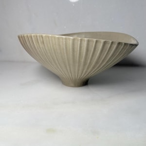 May include: A beige ceramic bowl with a wide, flared rim and a tapered base. The exterior features a series of vertical, recessed fluted lines that extend from the base to the top edge, creating a textured, ribbed appearance throughout the surface.