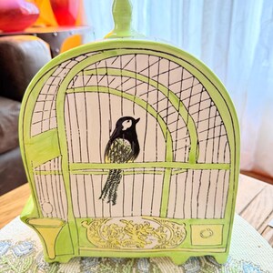 May include: A ceramic birdcage-shaped decorative piece in shades of green, white, and black. The cage design features a black and white bird inside. The base has a decorative floral design. The piece is sitting on a beaded placemat.
