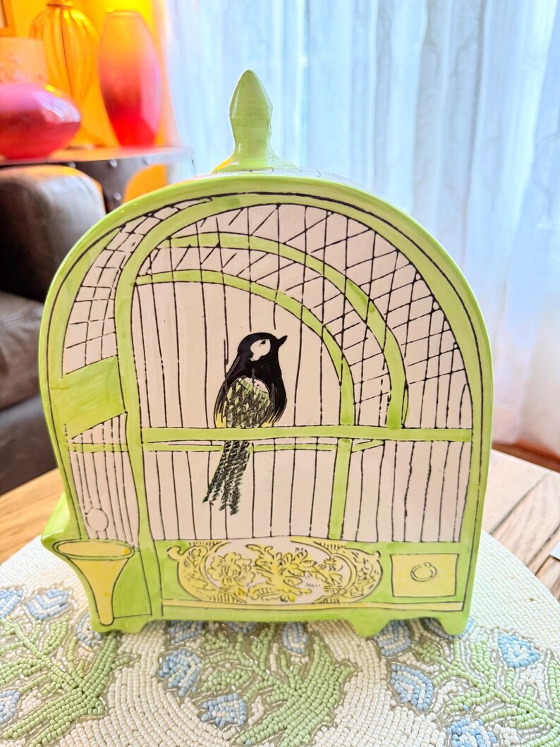 May include: A ceramic birdcage-shaped decorative piece in shades of green, white, and black. The cage design features a black and white bird inside. The base has a decorative floral design. The piece is sitting on a beaded placemat.