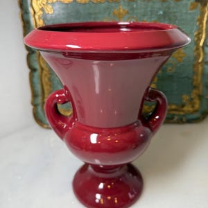 Pode incluir: Um vaso de cerâmica bordô brilhante com uma borda larga e flare e duas pequenas alças. O vaso tem uma base bulbosa e uma superfície lisa e polida. O fundo inclui uma bandeja decorativa.