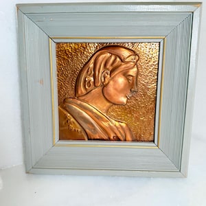 May include: A copper-toned relief sculpture of a woman in profile, framed in a light blue wooden frame with a gold inner edge. The artwork features detailed textures and a classical aesthetic, suitable for wall decor.