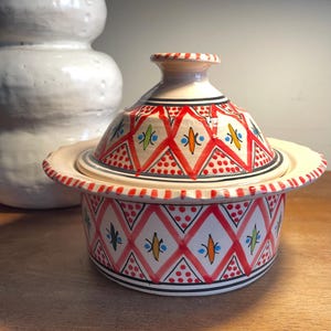 May include: A decorative ceramic tagine pot with a lid. The pot is white with a red and black geometric pattern. The lid has a knob and is decorated with red diamonds and colorful accents. The tagine sits on a wooden surface.
