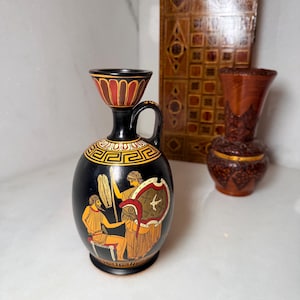 May include: A black ceramic lekythos vase featuring a painted scene of two figures in orange and white, with a Greek key pattern around the shoulder and a flared rim. The vase has a single handle and a narrow neck, showcasing detailed classical art.