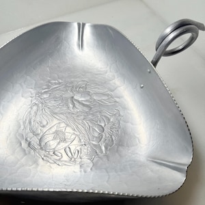 May include: A silver-colored, triangular serving dish with a floral embossed design in the center. The dish has a looped handle and a textured surface. The rim is scalloped, adding a decorative touch to this vintage serving piece.