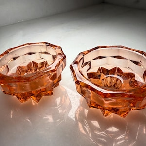 May include: Two vintage, faceted, orange-colored glass bowls. The bowls have a geometric design with a starburst pattern in the center. The bowls are likely used for serving or display.