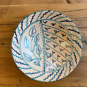 May include: A handmade ceramic plate with a cream-colored base and blue decorative elements. The plate features a leaf design on one side and a geometric pattern on the other. The rim is adorned with a wavy blue line.