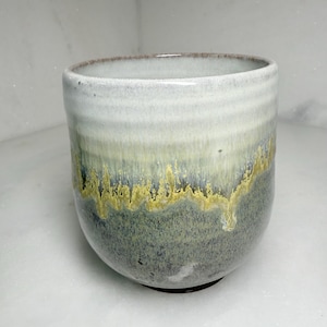 May include: A handmade ceramic cup with a unique glaze. The cup features a gradient of colors, starting with a light gray at the top, transitioning to green and yellow, and ending with a dark gray at the base. The cup has a rounded shape.