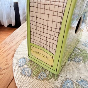 May include: A ceramic birdhouse with a green frame and a white grid pattern. The word "maison" is written on a small yellow rectangle. The birdhouse sits on a beaded placemat with blue and green floral designs.
