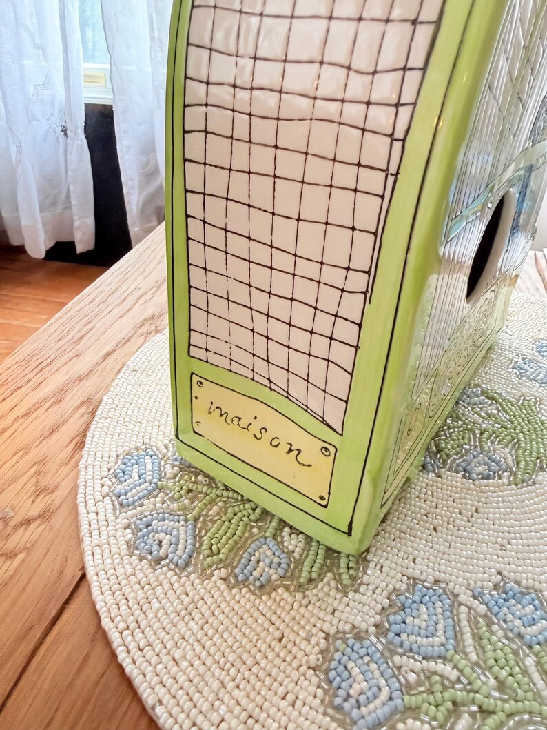 May include: A ceramic birdhouse with a green frame and a white grid pattern. The word "maison" is written on a small yellow rectangle. The birdhouse sits on a beaded placemat with blue and green floral designs.