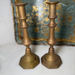 May include: Two antique brass candlesticks. Each candlestick has a hexagonal base and a slender, ornate stem. The candlesticks are a warm, golden color and are set against a blurred background of a teal tray with gold accents.