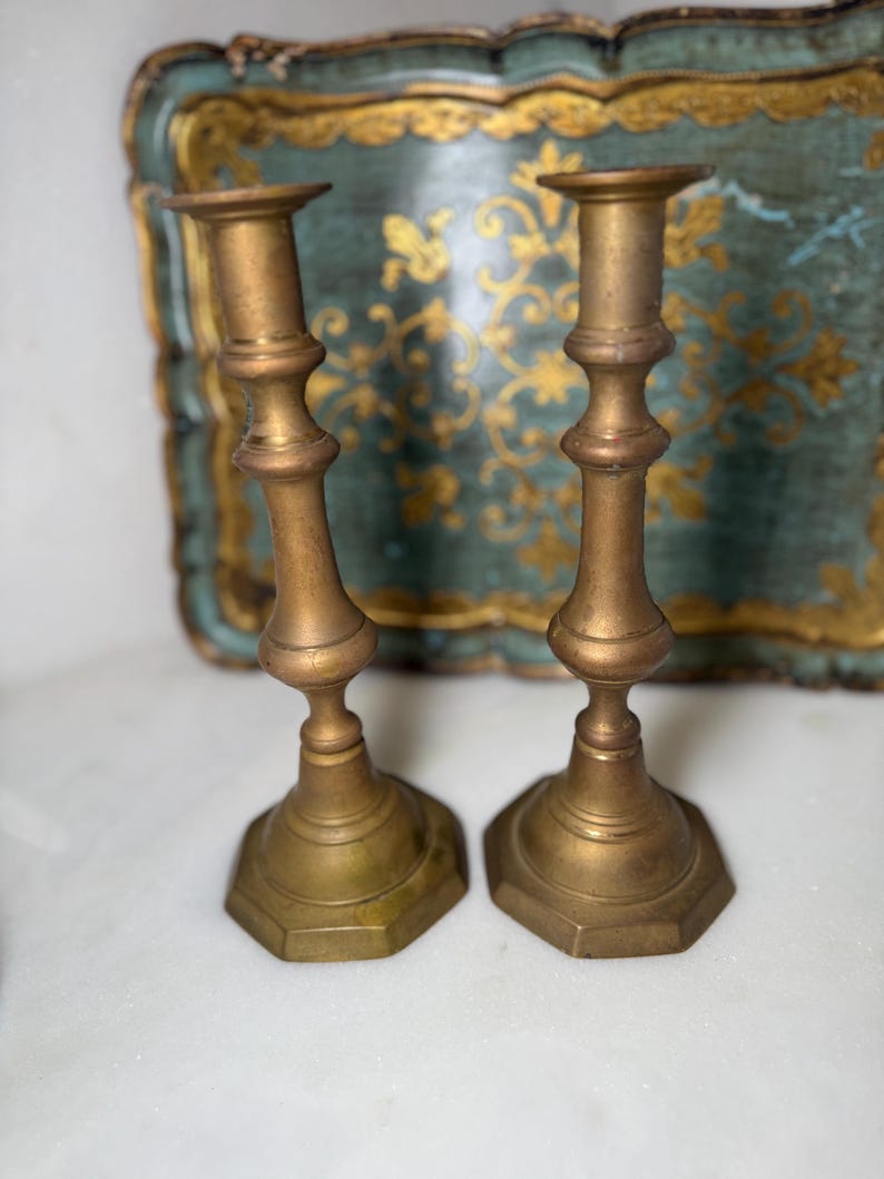 May include: Two antique brass candlesticks. Each candlestick has a hexagonal base and a slender, ornate stem. The candlesticks are a warm, golden color and are set against a blurred background of a teal tray with gold accents.
