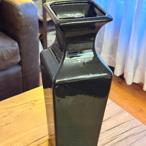 May include: A tall, black ceramic vase with a glossy finish. The vase has a square opening and a rectangular body that tapers slightly towards the base. The vase is sitting on a wooden surface.