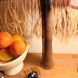 May include: A tall, slender ceramic vase with a gradient of brown hues, transitioning from dark at the top to lighter shades towards the base. The vase is displayed on a wooden surface, alongside a decorative glass turtle and a bowl of oranges.
