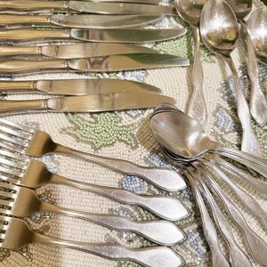 May include: A collection of vintage silverware, including knives, spoons, and forks, arranged on a beaded placemat. The silverware is made of a shiny, silver-colored metal. The placemat has a floral pattern with blue, green, and white beads.