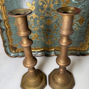 May include: Two antique brass candlesticks with a tarnished finish. Each candlestick has a hexagonal base, a bulbous midsection, and a cup-shaped top for holding candles. The background features a decorative tray with gold accents.