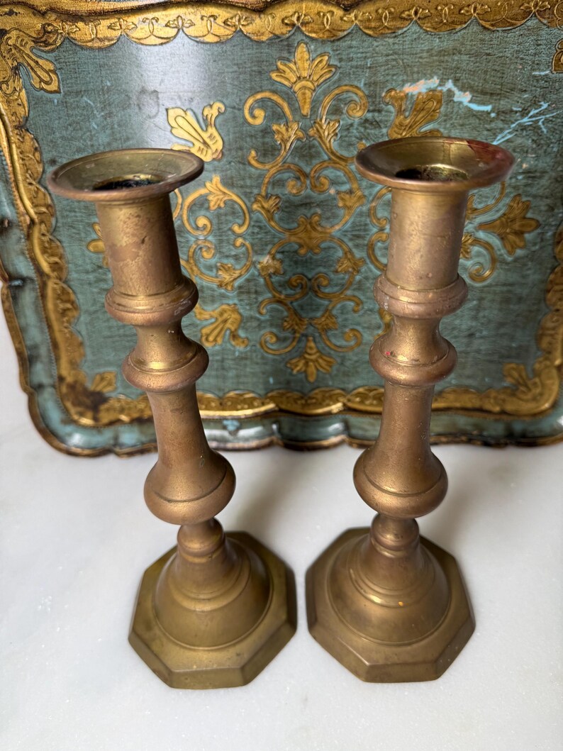 May include: Two antique brass candlesticks with a tarnished finish. Each candlestick has a hexagonal base, a bulbous midsection, and a cup-shaped top for holding candles. The background features a decorative tray with gold accents.