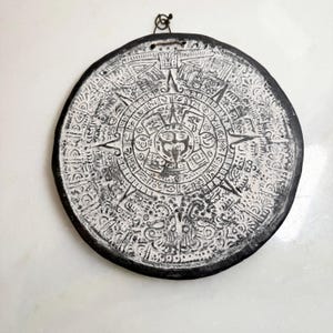 May include: A circular wall plaque featuring a detailed relief of the Aztec sun stone calendar. The piece is finished in a high-contrast black and white color scheme, with a central face surrounded by intricate geometric patterns and symbols.