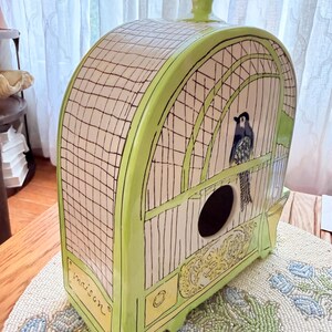 May include: A decorative birdhouse in a cage design. The ceramic birdhouse is primarily white with a green trim and features a black bird illustration. The word "maison" is written on the front. The birdhouse has a round opening and a decorative top.