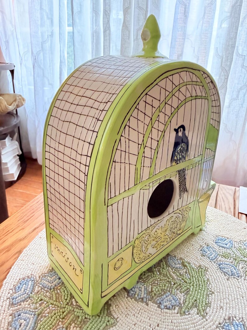 May include: A decorative birdhouse in a cage design. The ceramic birdhouse is primarily white with a green trim and features a black bird illustration. The word "maison" is written on the front. The birdhouse has a round opening and a decorative top.