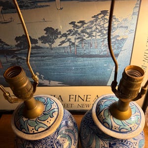 May include: Pair of ceramic table lamps with brass fixtures. The lamps feature a blue and white floral design with a light blue and green accent. The lamps are round and have a classic, vintage aesthetic.