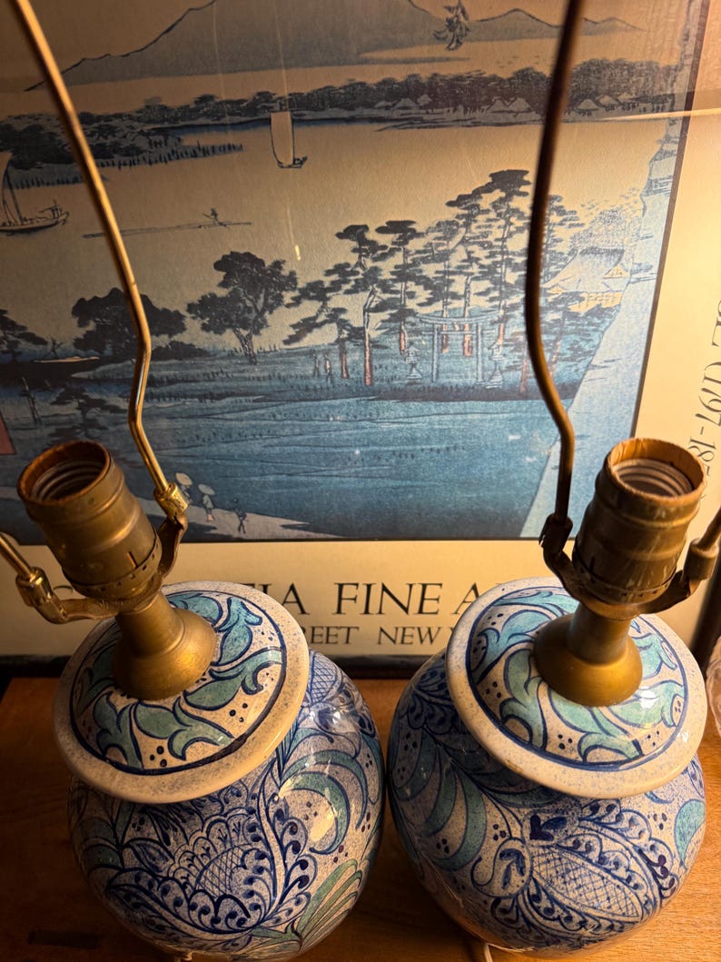 May include: Pair of ceramic table lamps with brass fixtures. The lamps feature a blue and white floral design with a light blue and green accent. The lamps are round and have a classic, vintage aesthetic.