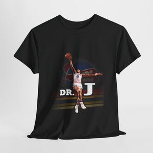 May include: Black t-shirt featuring a graphic of a basketball player in mid-air, holding a basketball. The design includes the text "DR. J" in white. The player is wearing a white and red uniform. The background has a basketball and stripes.
