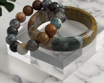 May include: Ocean Jasper bangle and bracelet set. The set includes a 60 mm bangle and a bracelet with 12 mm beads. The bangle is a mix of brown, green, and tan colors. The bracelet features a variety of earth-toned beads. The set is displayed on a clear acrylic stand.
