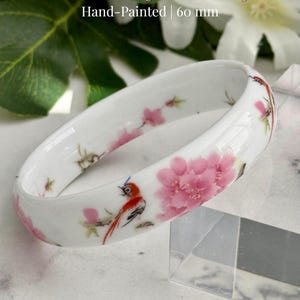 May include: A white porcelain bangle with hand-painted cherry blossom and bird designs. The bangle features pink flowers and red birds, with a diameter of 60 mm.