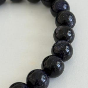 May include: A bracelet composed of round, polished dark blue sandstone beads. Each bead features a smooth, glossy surface with embedded reflective metallic flecks that create a shimmering, star-like appearance throughout the deep blue material.