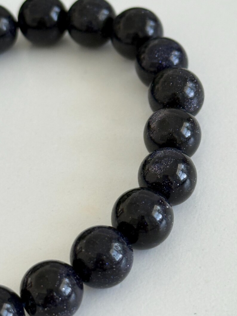 May include: A bracelet composed of round, polished dark blue sandstone beads. Each bead features a smooth, glossy surface with embedded reflective metallic flecks that create a shimmering, star-like appearance throughout the deep blue material.