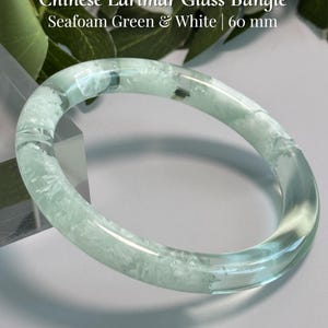 May include: A seafoam green and white Chinese Larimar glass bangle with a 60 mm diameter. The text on the image reads: Chinese Larimar Glass Bangle, Seafoam Green & White | 60 mm, and AmorGemsBoutique. The bangle has a smooth, translucent finish.