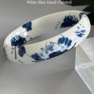 May include: A white porcelain bangle bracelet featuring hand-painted blue floral patterns. The text Porcelain Floral Bangle White Blue Hand-Painted and AmorGemsBoutique is visible. The bangle has a smooth, glossy finish and a rounded, continuous circular shape.