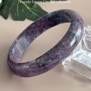May include: A polished bangle made of ruby zoisite featuring mottled purple and green coloration. The bangle has a smooth, rounded surface and a diameter of 58.5 mm. Text on the image reads Ruby Zoisite Bangle, Purple Green Crystal, 58.5 mm.