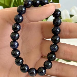 May include: A stretch bracelet composed of round, dark blue sandstone beads. Each bead features a polished, reflective surface with embedded glittery particles that create a sparkling effect resembling a starry night sky, strung on an elastic cord.