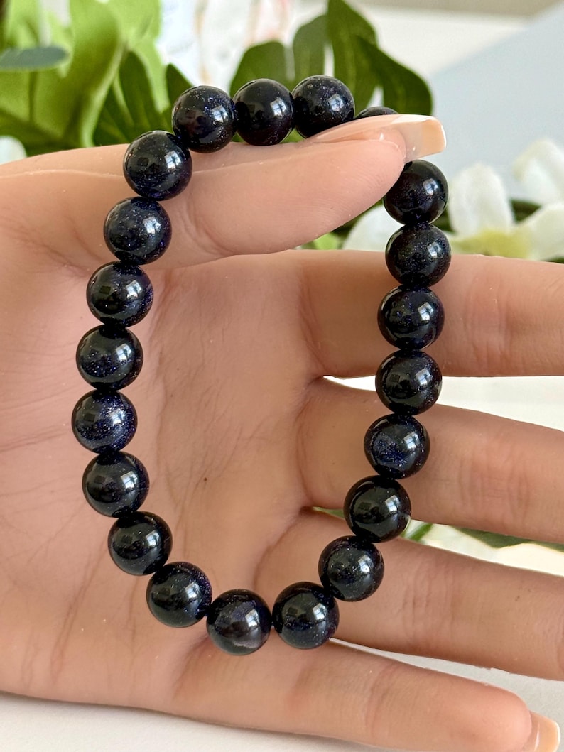 May include: A stretch bracelet composed of round, dark blue sandstone beads. Each bead features a polished, reflective surface with embedded glittery particles that create a sparkling effect resembling a starry night sky, strung on an elastic cord.