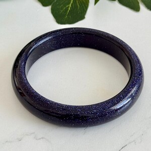 May include: A smooth, circular bangle bracelet crafted from dark blue sandstone, featuring a polished finish with numerous tiny, reflective silver-toned glitter particles embedded throughout the material, creating a starry night sky appearance.