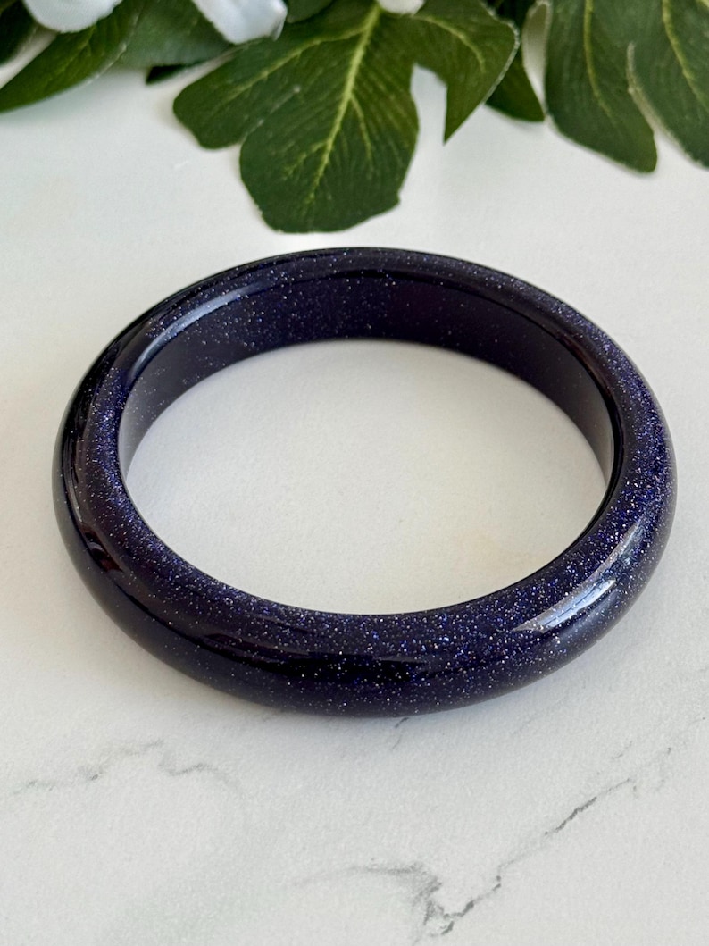 May include: A smooth, circular bangle bracelet crafted from dark blue sandstone, featuring a polished finish with numerous tiny, reflective silver-toned glitter particles embedded throughout the material, creating a starry night sky appearance.