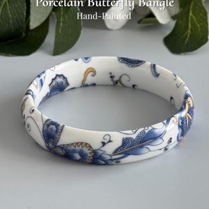 May include: A white porcelain bangle bracelet featuring a hand-painted blue and gold butterfly and floral pattern. The text Porcelain Butterfly Bangle Hand-Painted and AmorGemsBoutique is visible. The bangle has a smooth, glossy finish and a circular shape.