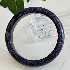 May include: A circular bangle bracelet crafted from dark blue sandstone, featuring a smooth, polished surface embedded with numerous tiny, reflective glitter-like particles that create a shimmering, star-filled appearance throughout the material.