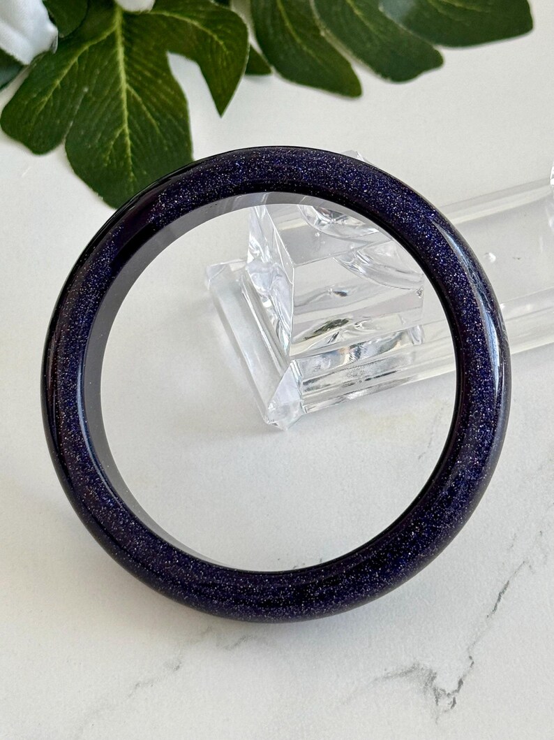 May include: A circular bangle bracelet crafted from dark blue sandstone, featuring a smooth, polished surface embedded with numerous tiny, reflective glitter-like particles that create a shimmering, star-filled appearance throughout the material.