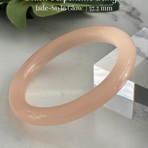 May include: A blush-colored serpentine bangle with a jade-style glow. The text on the image reads "Blush Serpentine Bangle" and "Jade-Style Glow | 57.2 mm". The bangle is a light pink color and is approximately 2.25 inches in diameter.