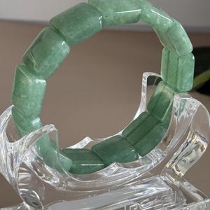 Pu&ograve; includere: Un braccialetto di giada verde composto da perline rettangolari. Il braccialetto &egrave; esposto su un supporto decorativo trasparente. La giada ha una qualit&agrave; traslucida e le perline sono lucidate. Il supporto ha un design ornato.