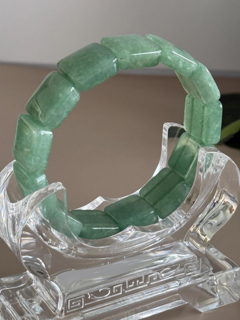 Pu&ograve; includere: Un braccialetto di giada verde composto da perline rettangolari. Il braccialetto &egrave; esposto su un supporto decorativo trasparente. La giada ha una qualit&agrave; traslucida e le perline sono lucidate. Il supporto ha un design ornato.