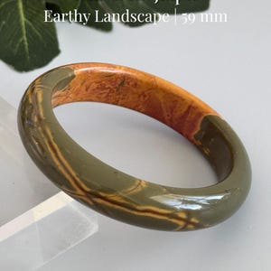 May include: A polished Red Creek Jasper bangle bracelet featuring a smooth, rounded surface with earthy tones of olive green, tan, and reddish-brown. The stone displays natural landscape-like patterns and has an inner diameter of 59 mm.