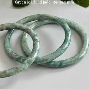 May include: Three green marbled jadeite bangles. The text on the image reads "Grade A Jadeite Green Marbled Jade | 59-59.3 mm". The bangles are a light green color with darker green marbling. The brand name "AmorGems Boutique" is also visible.