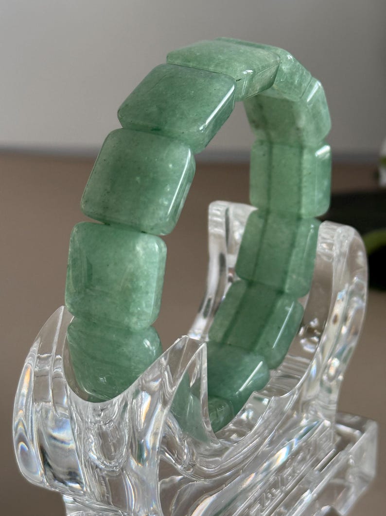 Pu&ograve; includere: Un braccialetto di avventurina verde con perline quadrate. Il braccialetto &egrave; esposto su un supporto trasparente e decorativo. Le perline hanno una superficie lucida e liscia e un aspetto traslucido. Il braccialetto &egrave; un gioiello.
