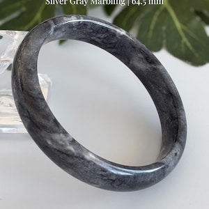 May include: A natural jade bangle with silver gray marbling, featuring a 64.5 mm diameter. The text Natural Jade Bangle, Silver Gray Marbling | 64.5 mm, and AmorGemsBoutique™ are visible on the image, showcasing a smooth, polished stone surface.